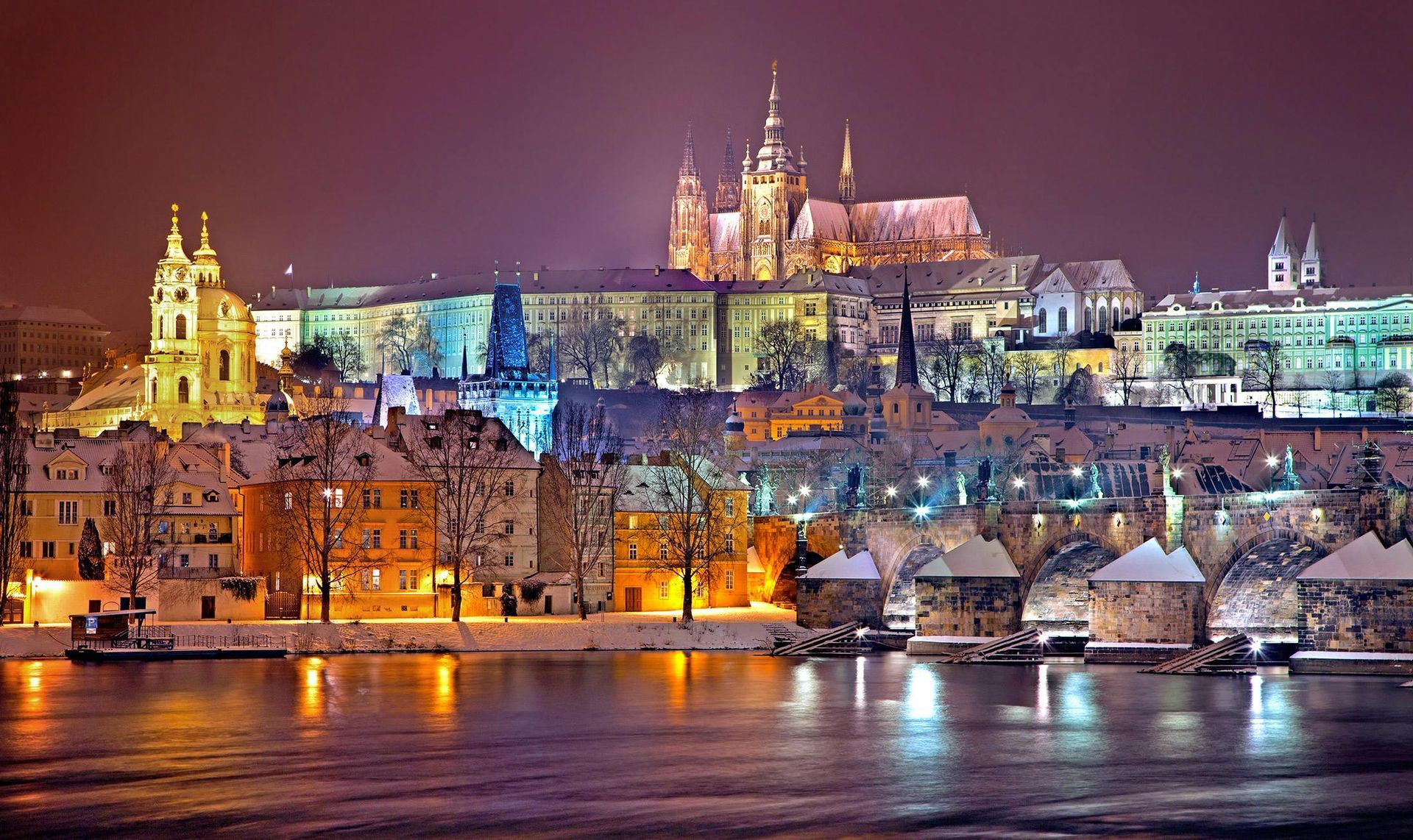 A city at night with a castle in the background and a river in the foreground.