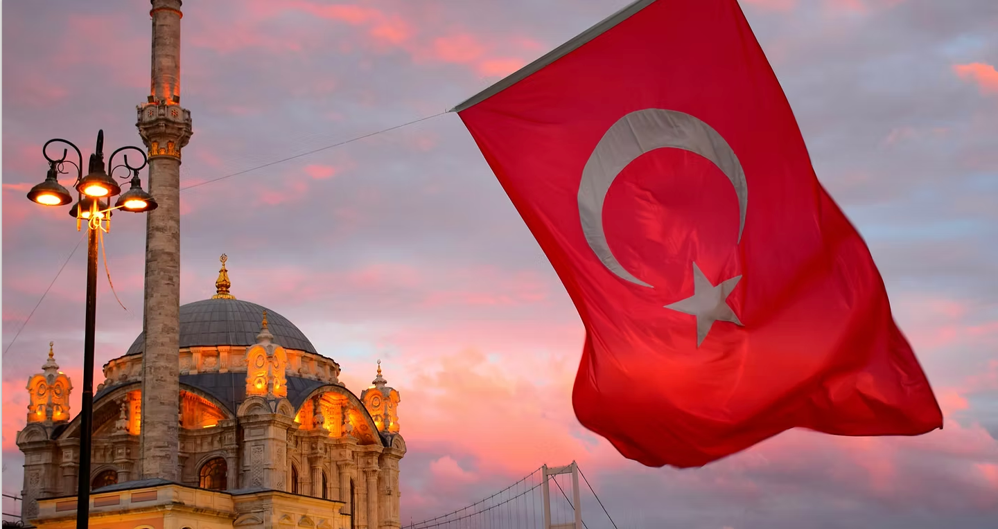 A red flag with a crescent moon and star is flying in front of a mosque.