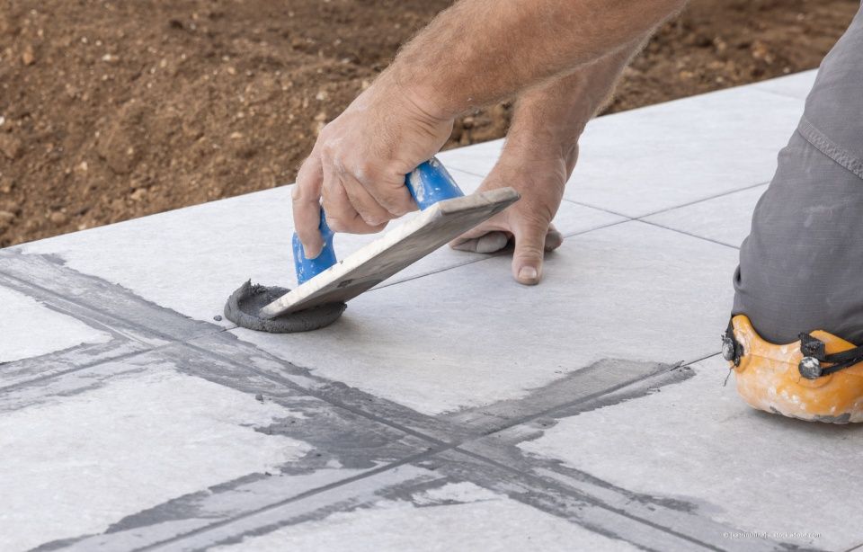 Person, die im Freien mit einer Kelle Fugenmörtel zwischen grauen Fliesen aufträgt.