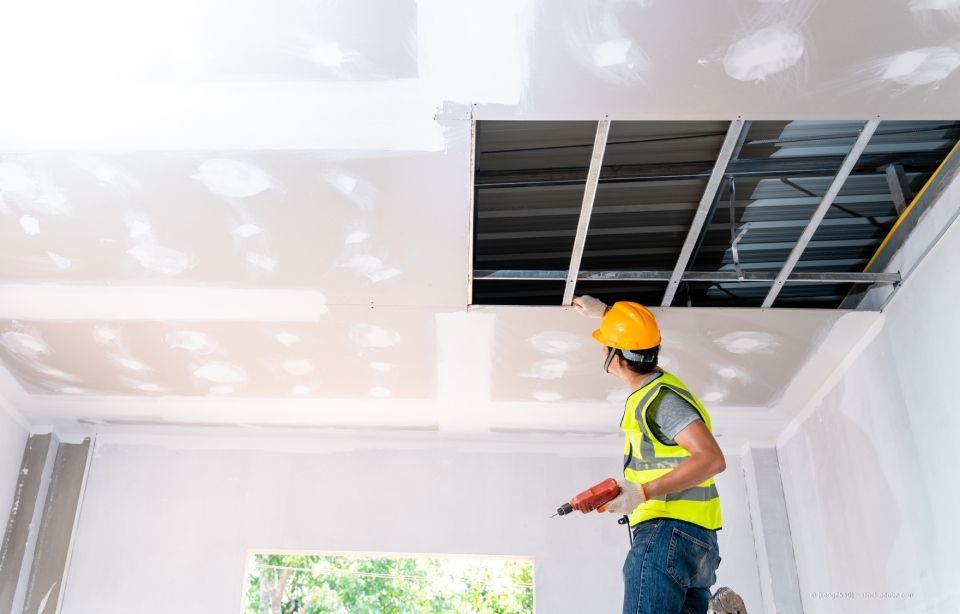 Bauarbeiter in gelber Weste und Schutzhelm installiert Trockenbauwände an einer Decke.