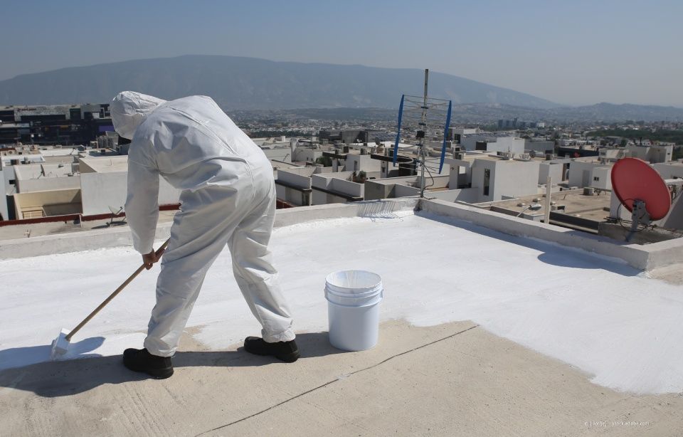 Person im weißen Schutzanzug bemalt ein weißes Dach, Eimer in der Nähe, Stadtbild im Hintergrund.