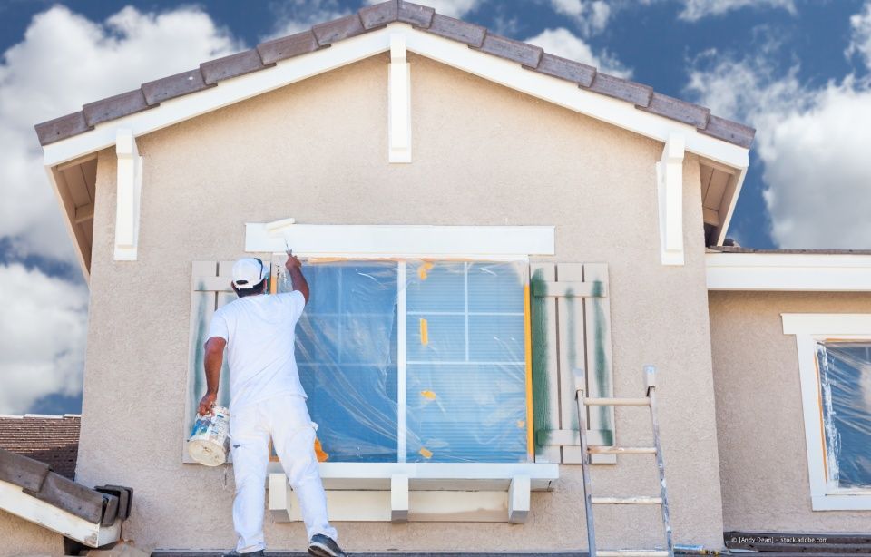 Person in Weiß streicht die äußere Fensterverkleidung eines beigen Hauses.