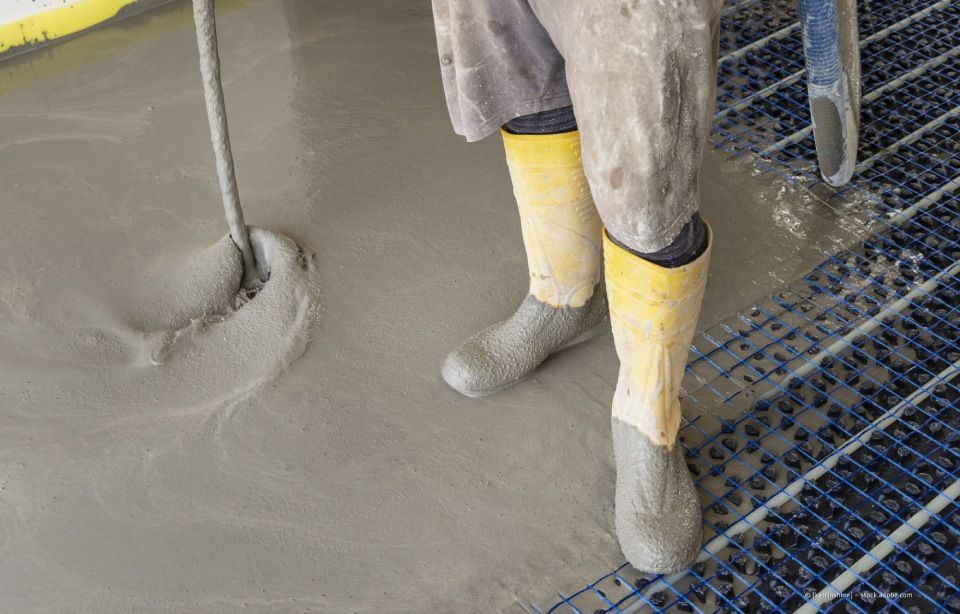 Person in Stiefeln gießt nassen Beton über blaue Fußbodenheizungsrohre.