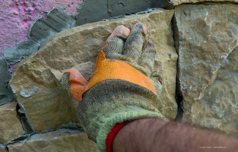 Behandschuhte Hand drückt einen hellbraunen Stein auf eine Wand, die gerade mit Mörtel errichtet wird.
