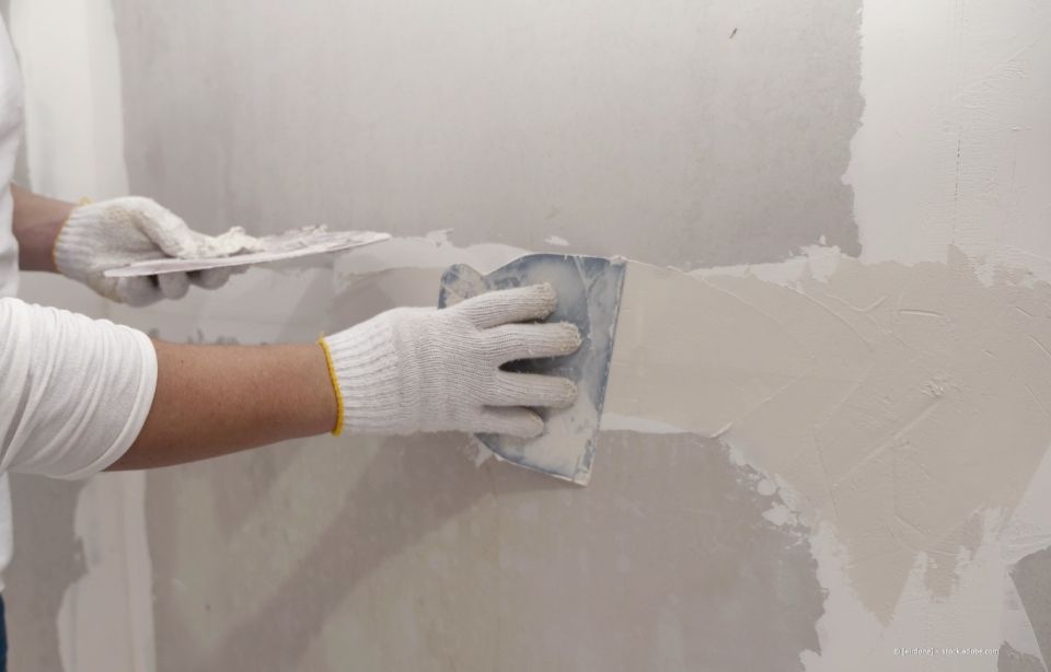 Person, die mit weißen Handschuhen Trockenbauspachtelmasse mit einem Spachtel auf eine Wand aufträgt.
