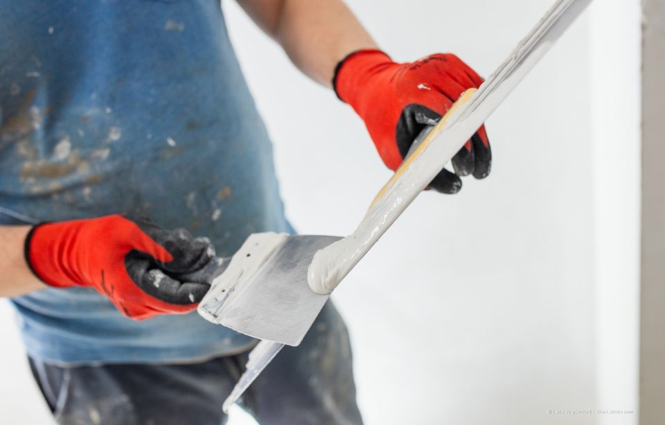 Eine Person mit roten Handschuhen glättet mit einem Spachtel die weiße Wandmasse.