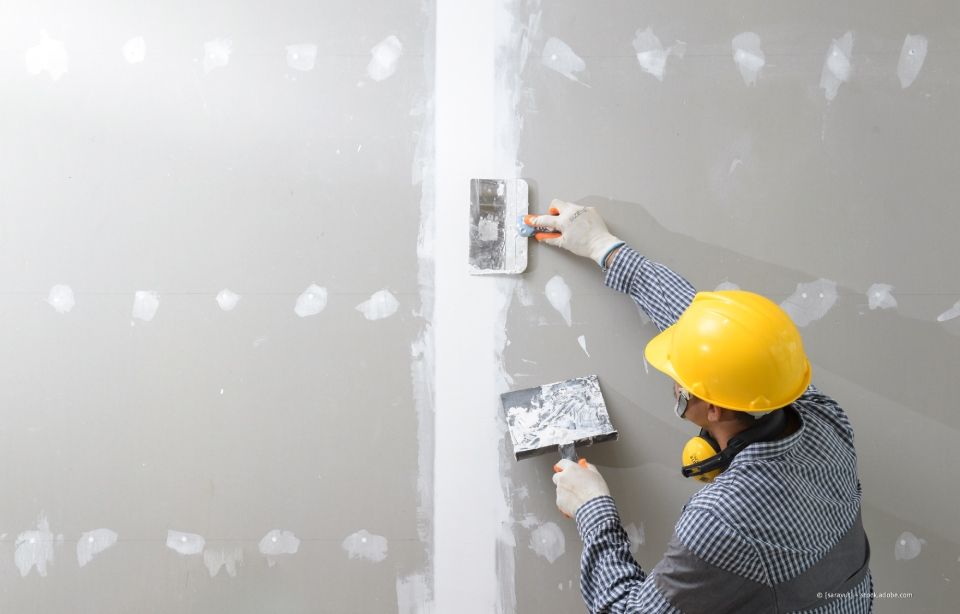 Person mit gelbem Schutzhelm und Handschuhen. Tragen Sie Trockenbauspachtelmasse auf eine Wand auf.