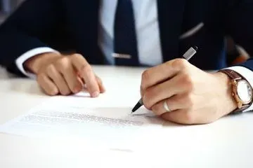 Una persona con traje oscuro, corbata y reloj se sienta en un escritorio blanco, sosteniendo un bolígrafo para firmar un documento.