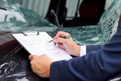 Un ajustador de seguros con un traje azul escribe en un portapapeles, de pie frente a un automóvil con el parabrisas destrozado.