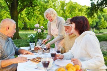 Cuatro personas disfrutan de un picnic al aire libre en una mesa con bebidas y comida en un parque soleado y verde.