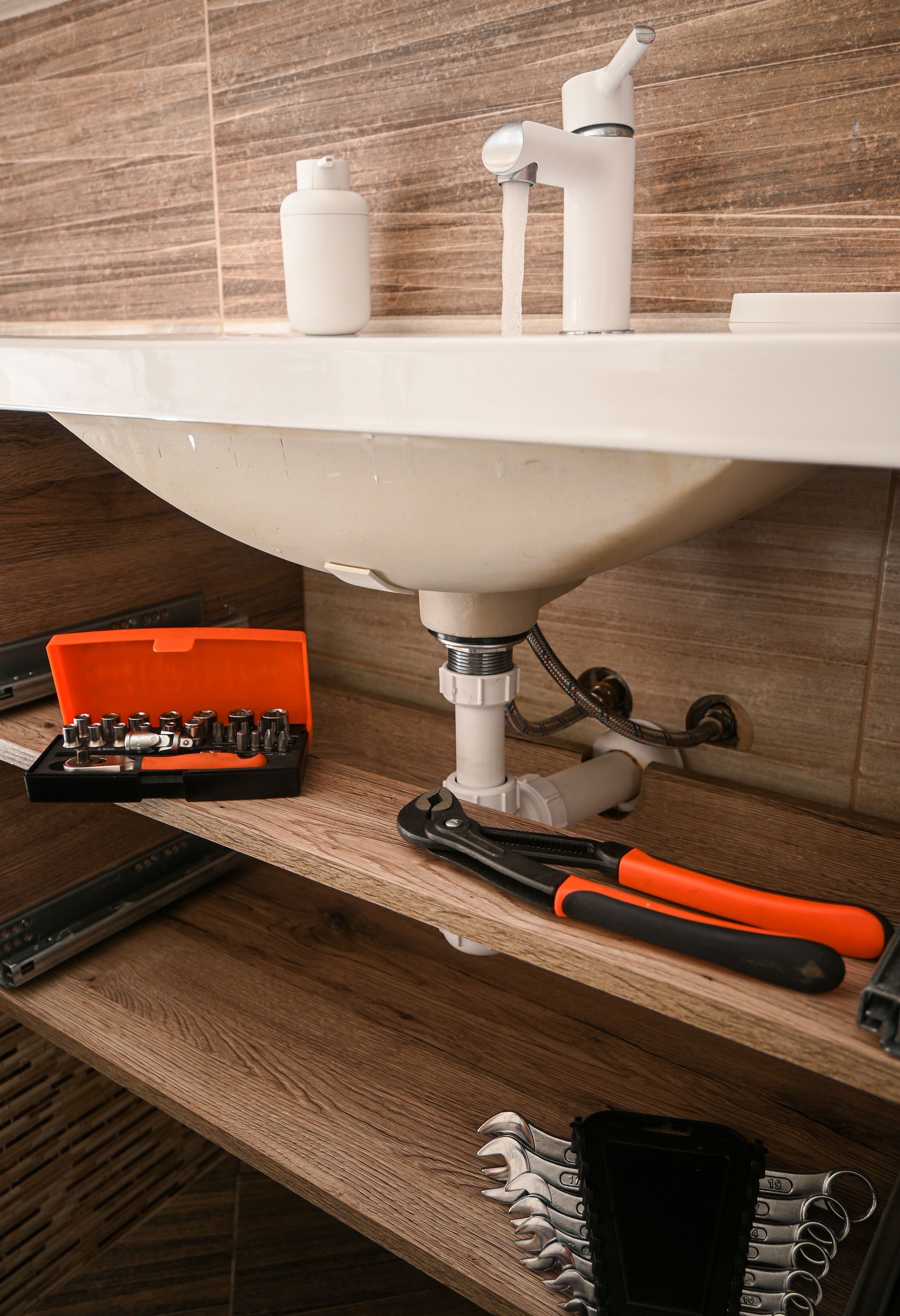 Lavabo de salle de bains avec tuyauterie apparente, outils de nettoyage et clé à pipe sous l'évier.