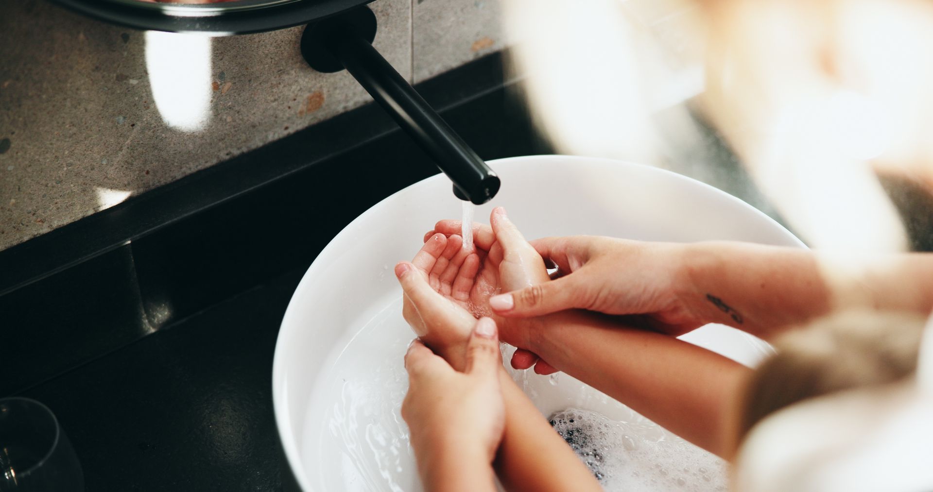 Deux personnes qui se lavent les mains sous l'eau courante d'un robinet dans un lavabo blanc rempli de mousse de savon.