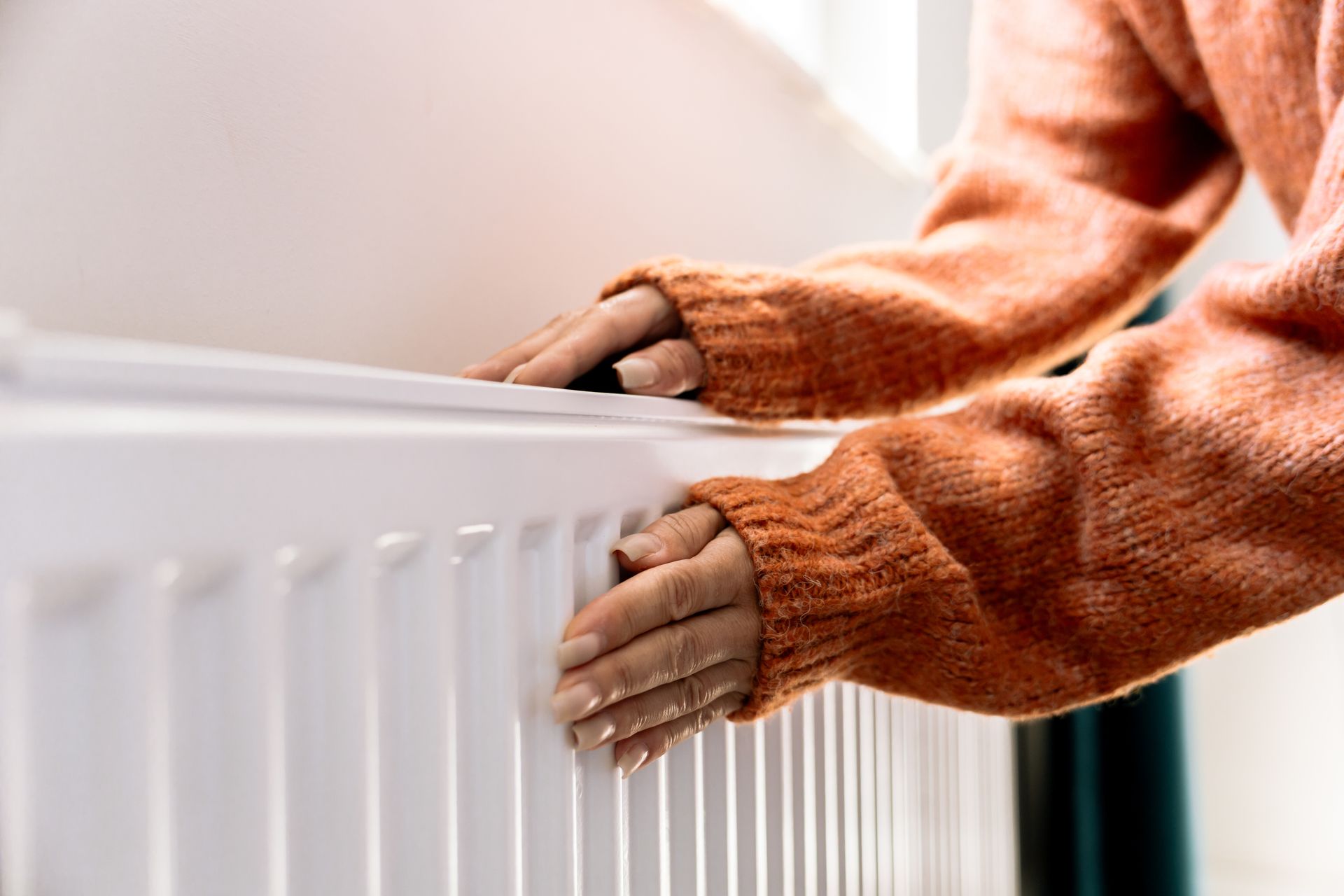 Mains féminines se réchauffent sur un radiateur blanc, vêtu d'un pull orange.