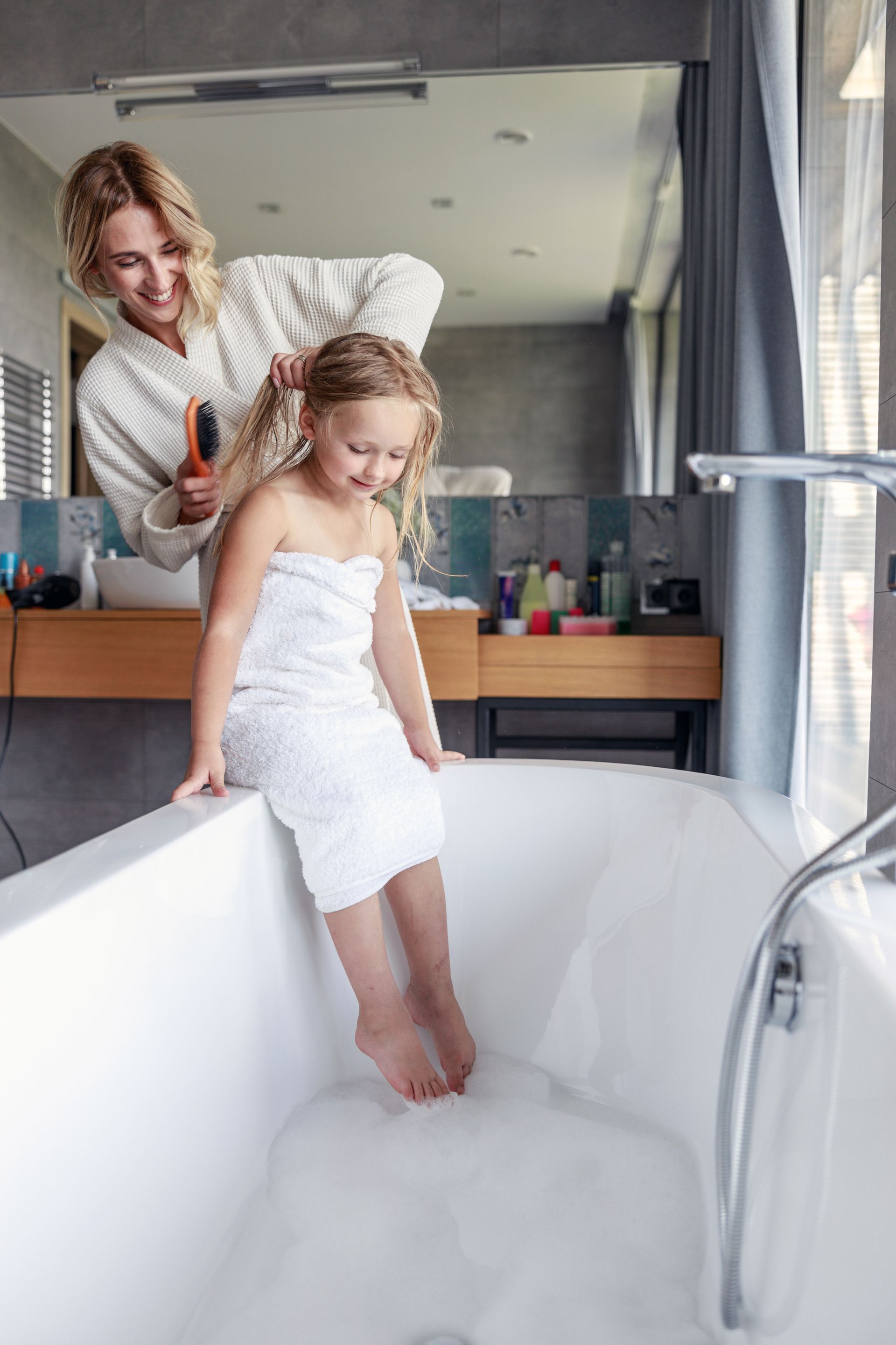 Une femme coiffe une petite fille assise sur le rebord d'une baignoire blanche dans une salle de bains.