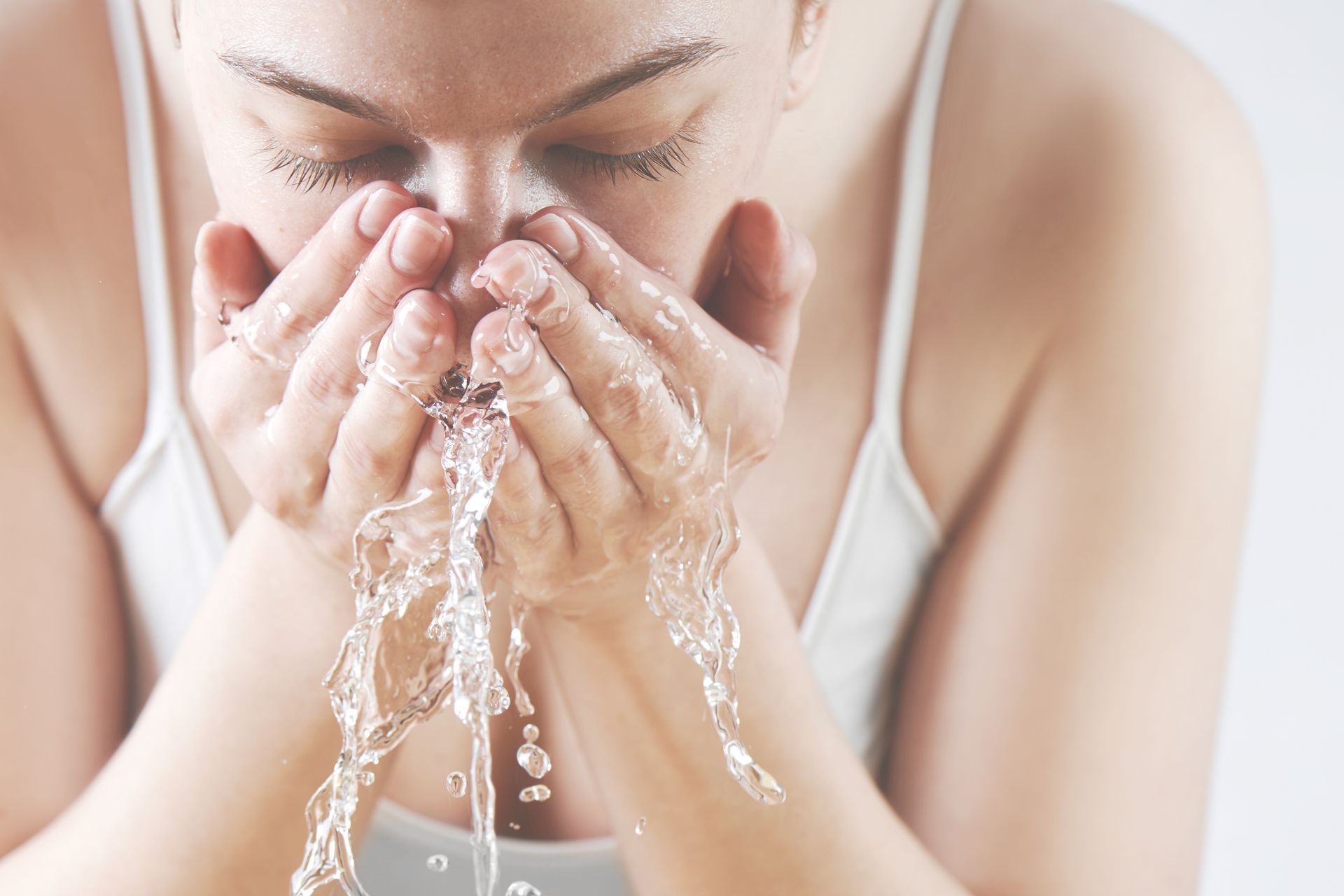 Femme se lavant le visage à l'eau au-dessus d'un lavabo, vêtue d'un débardeur blanc.