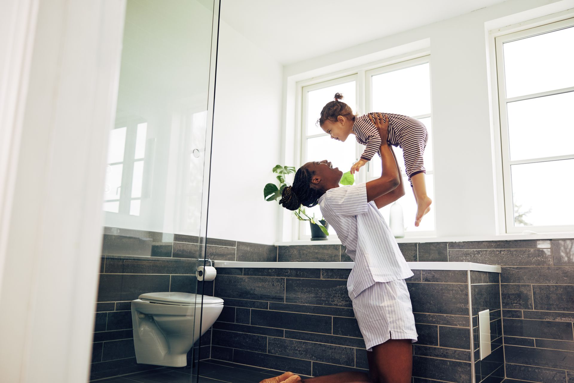 Un adulte soulève un enfant dans une salle de bains lumineuse, à côté d'une baignoire et d'un mur carrelé foncé.