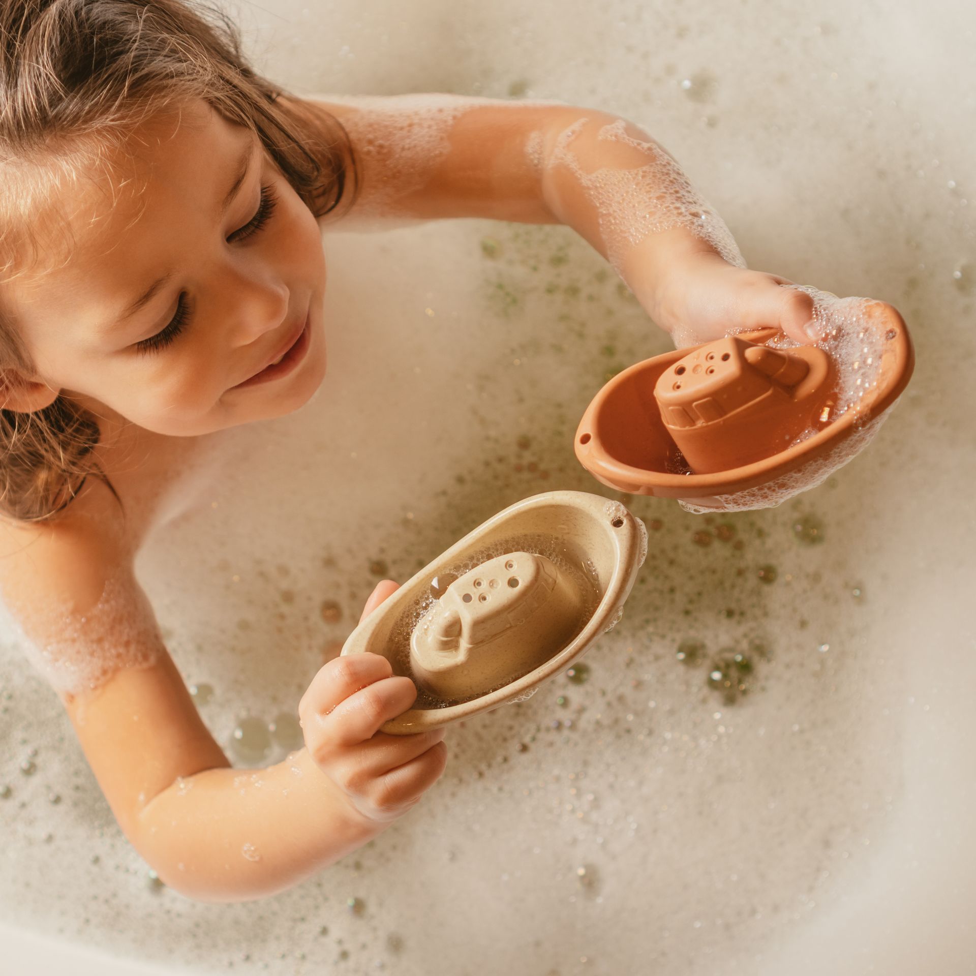 Enfant jouant dans un bain moussant, souriant en tenant deux petits bateaux.