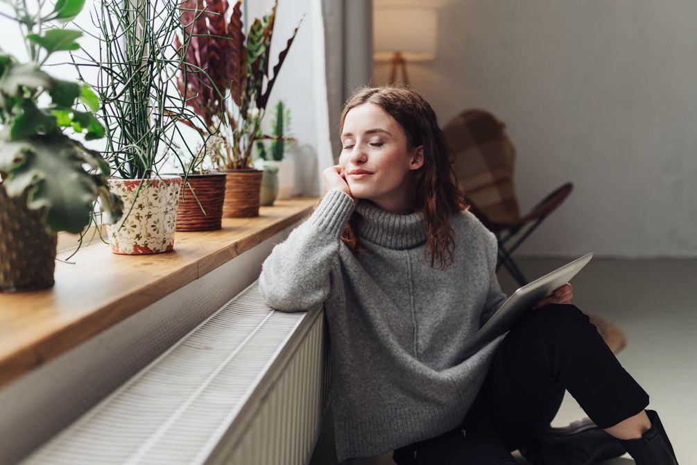 Une femme en pull gris est assise près d'une fenêtre, la tête appuyée contre un mur, en train de lire à côté de plantes en pot.