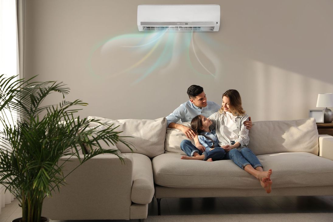 Une famille assise sur un canapé beige sous un climatiseur diffusant un flux d'air bleu frais dans un salon lumineux.