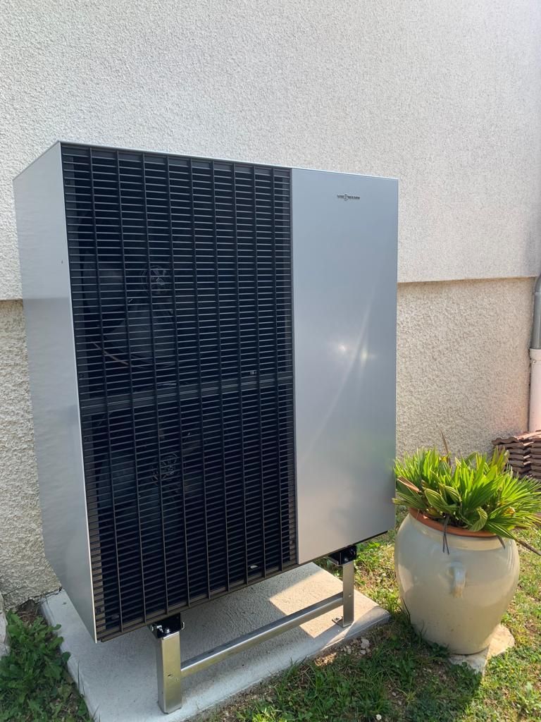 Grande unité de climatisation extérieure grise à côté d'un mur beige et d'une plante en pot au soleil.