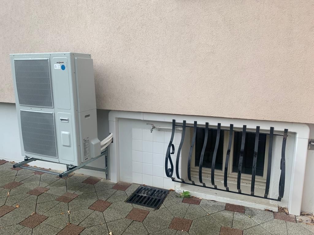 Unité de climatisation extérieure à côté d'une bouche d'aération murale avec câbles et tuyauterie apparents sur une terrasse pavée.