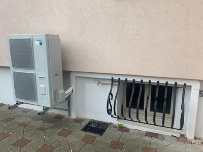 Unité de climatisation extérieure à côté d'une bouche d'aération murale avec câbles et tuyauterie apparents sur une terrasse pavée.