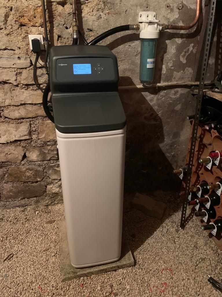 Système d'adoucisseur d'eau installé contre un mur de briques, avec tuyaux et vannes, dans une buanderie au sous-sol.