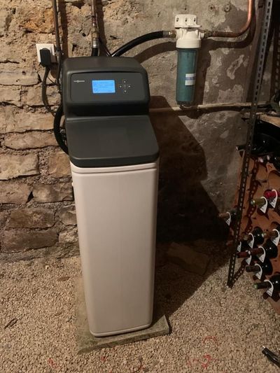 Système d'adoucisseur d'eau installé contre un mur de briques, avec tuyaux et vannes, dans une buanderie au sous-sol.