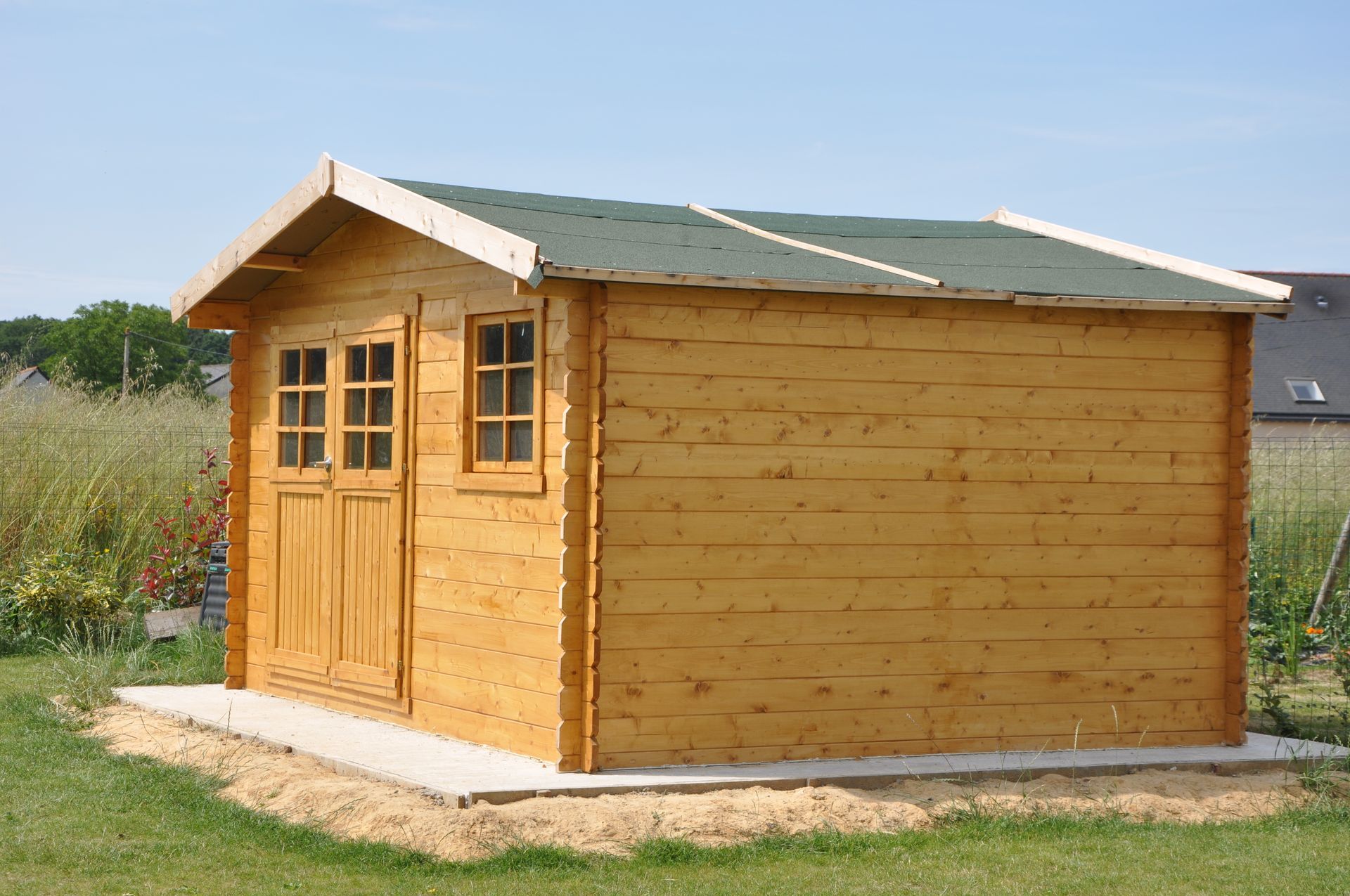 Abri de jardin en bois avec toit végétalisé, deux portes et une fenêtre, sur une base en béton.