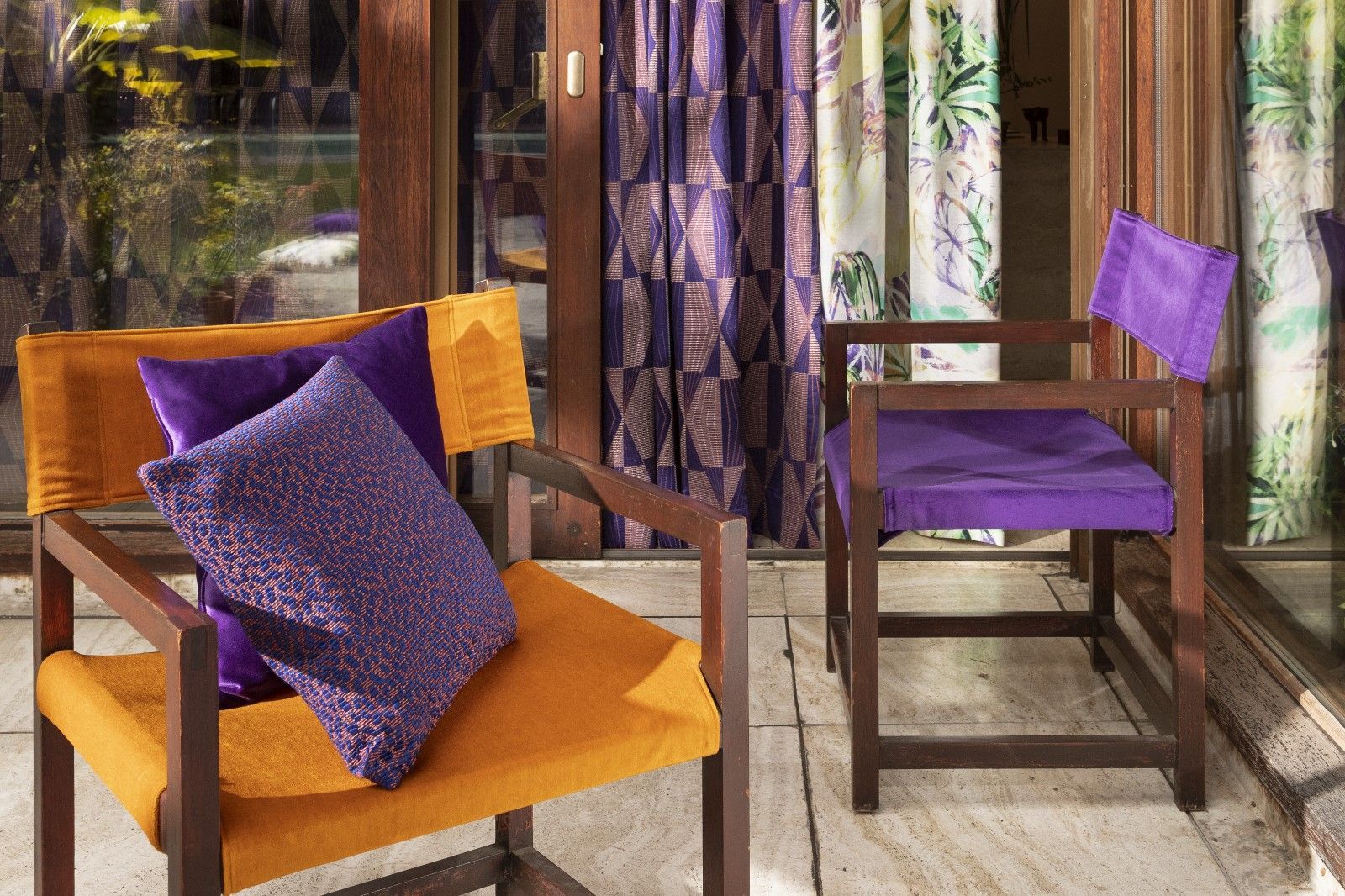 Zwei Holzstühle mit orangefarbenen und violetten Kissen, im Hintergrund Vorhänge.