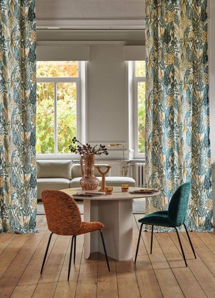 Esszimmer mit gemusterten Vorhängen, rundem Tisch, zwei Stühlen und Blick in ein Wohnzimmer mit Sofa.