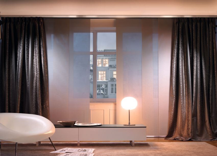 Wohnzimmer mit transparenten weißen Stoffbahnen, dunklen, glitzernden Vorhängen, moderner Lampe und Fensterblick.