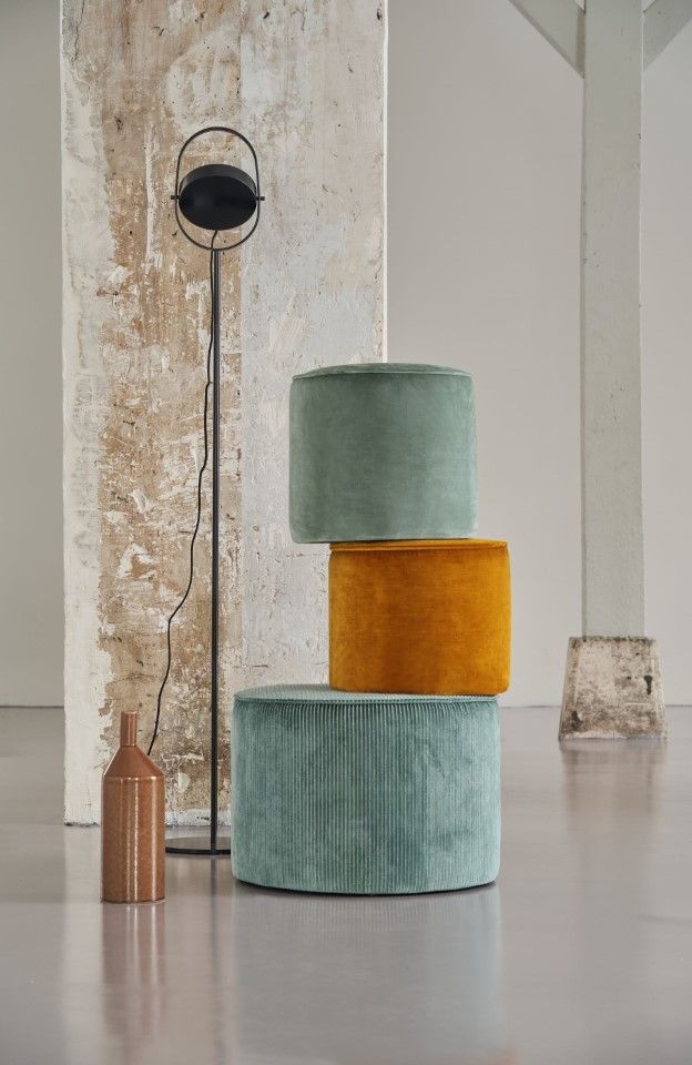Drei zylindrische Samt-Ottomane in Türkis und Gold, gestapelt vor einer strukturierten Wand.