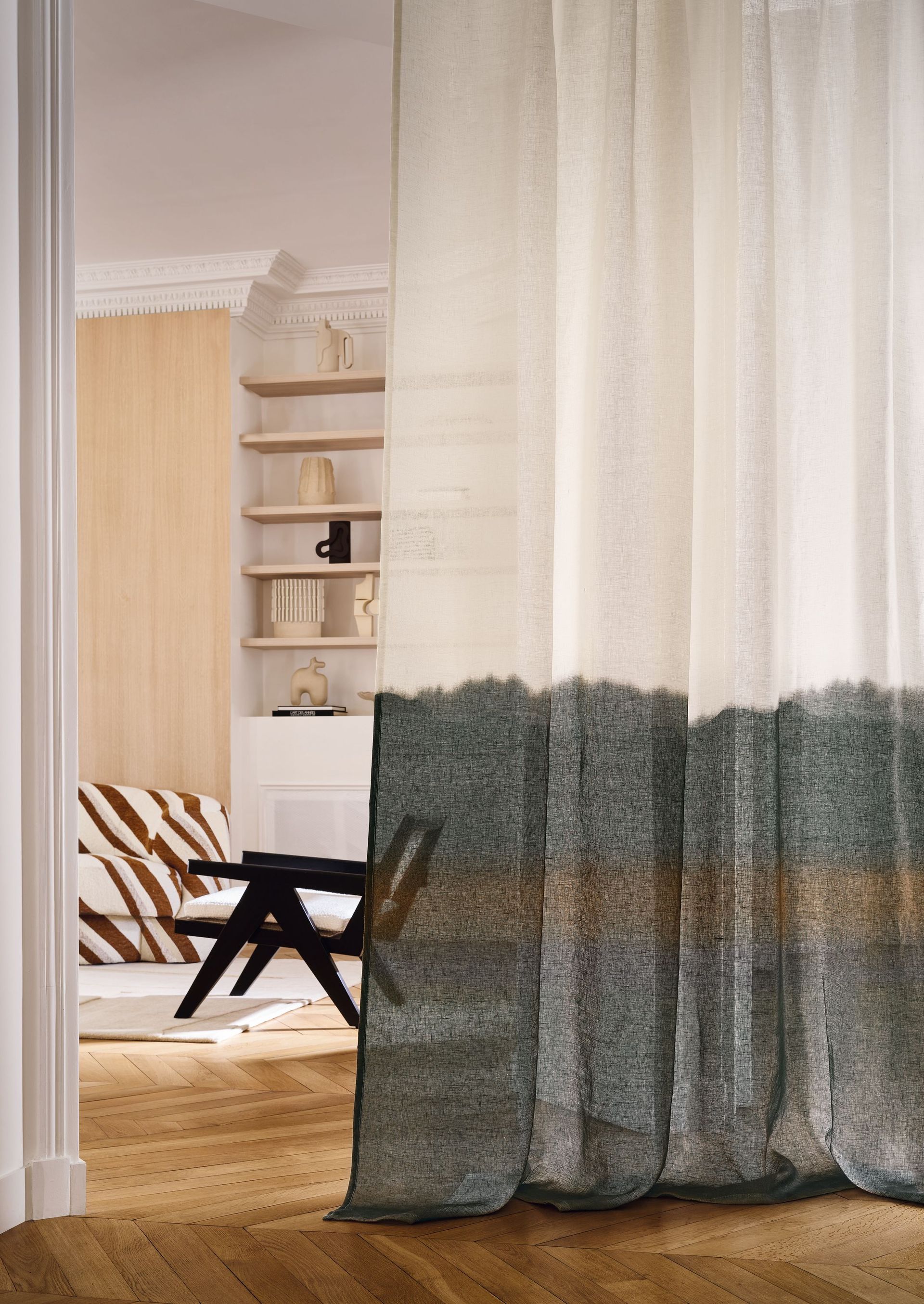 Transparenter Vorhang mit weißem Oberteil und grün-braunem Farbverlauf am unteren Rand, Blick durch die Tür in ein Zimmer mit Möbeln und Regalen.