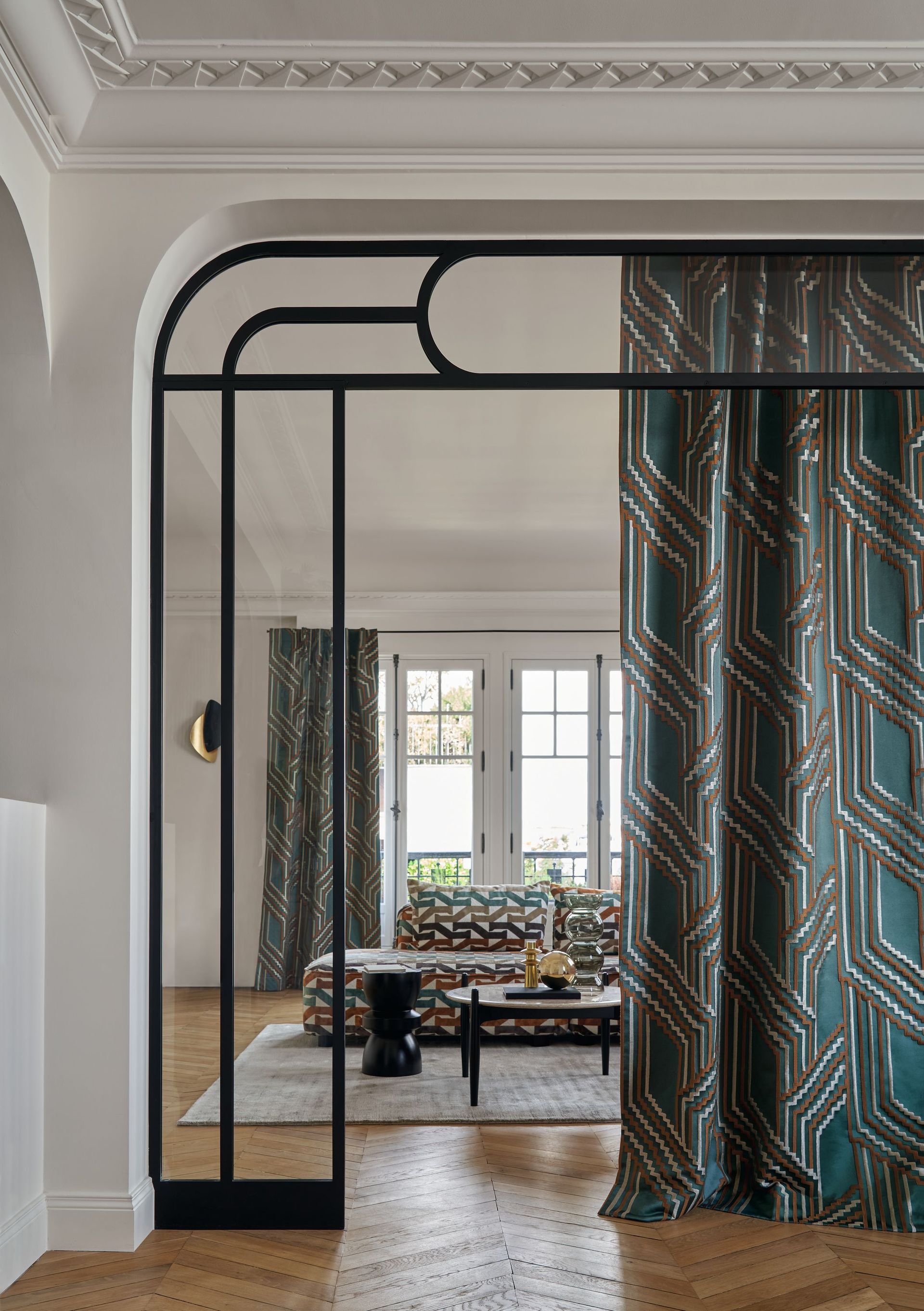 Elegantes Zimmer mit türkisfarbenen, geometrisch gemusterten Vorhängen, schwarzem Metalltürrahmen und gemustertem Sofa.