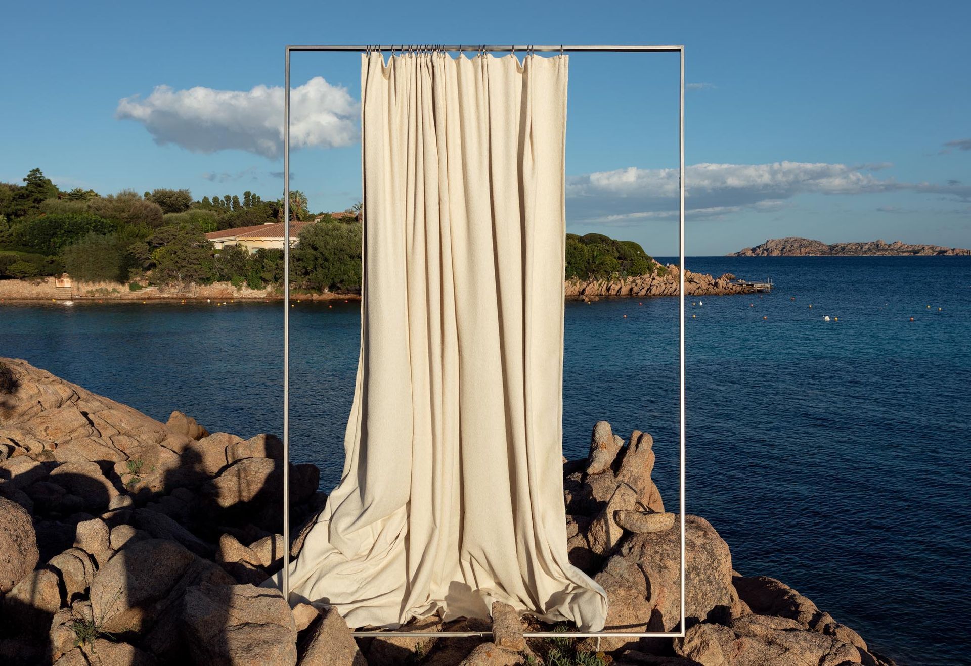 Cremefarbener Vorhang, der in einem Metallrahmen an einem felsigen Ufer hängt, im Hintergrund Meer und blauer Himmel.