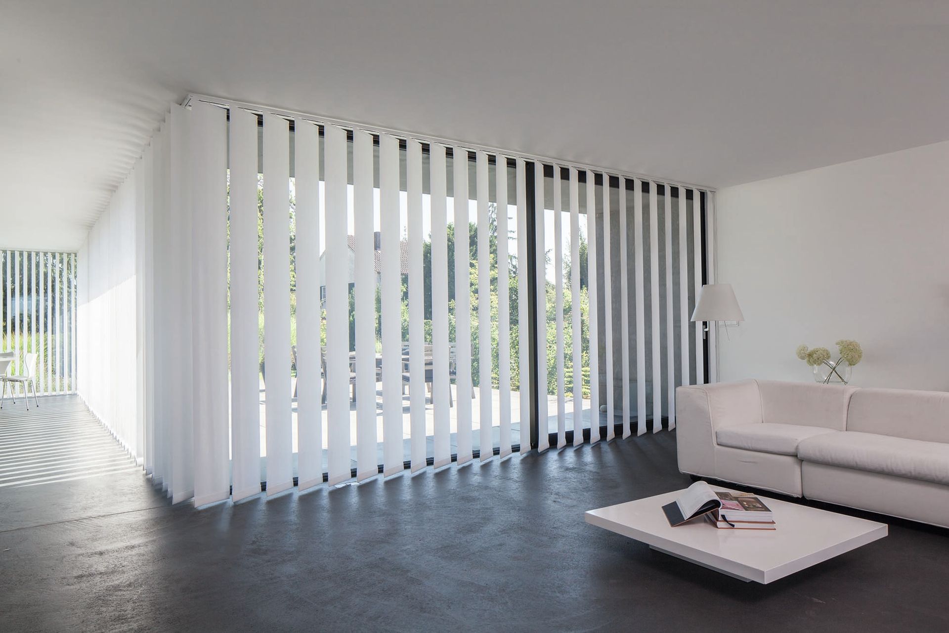 Weiße Vertikaljalousien verdecken ein großes Fenster in einem modernen, minimalistischen Wohnzimmer mit weißen Möbeln.