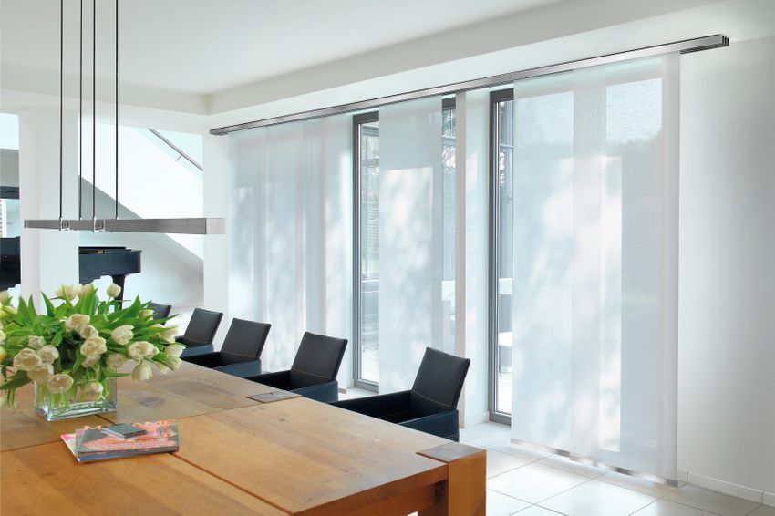 Esszimmer mit transparenten, weißen Flächenvorhängen an den Fenstern. Holztisch, schwarze Stühle, Blumen.