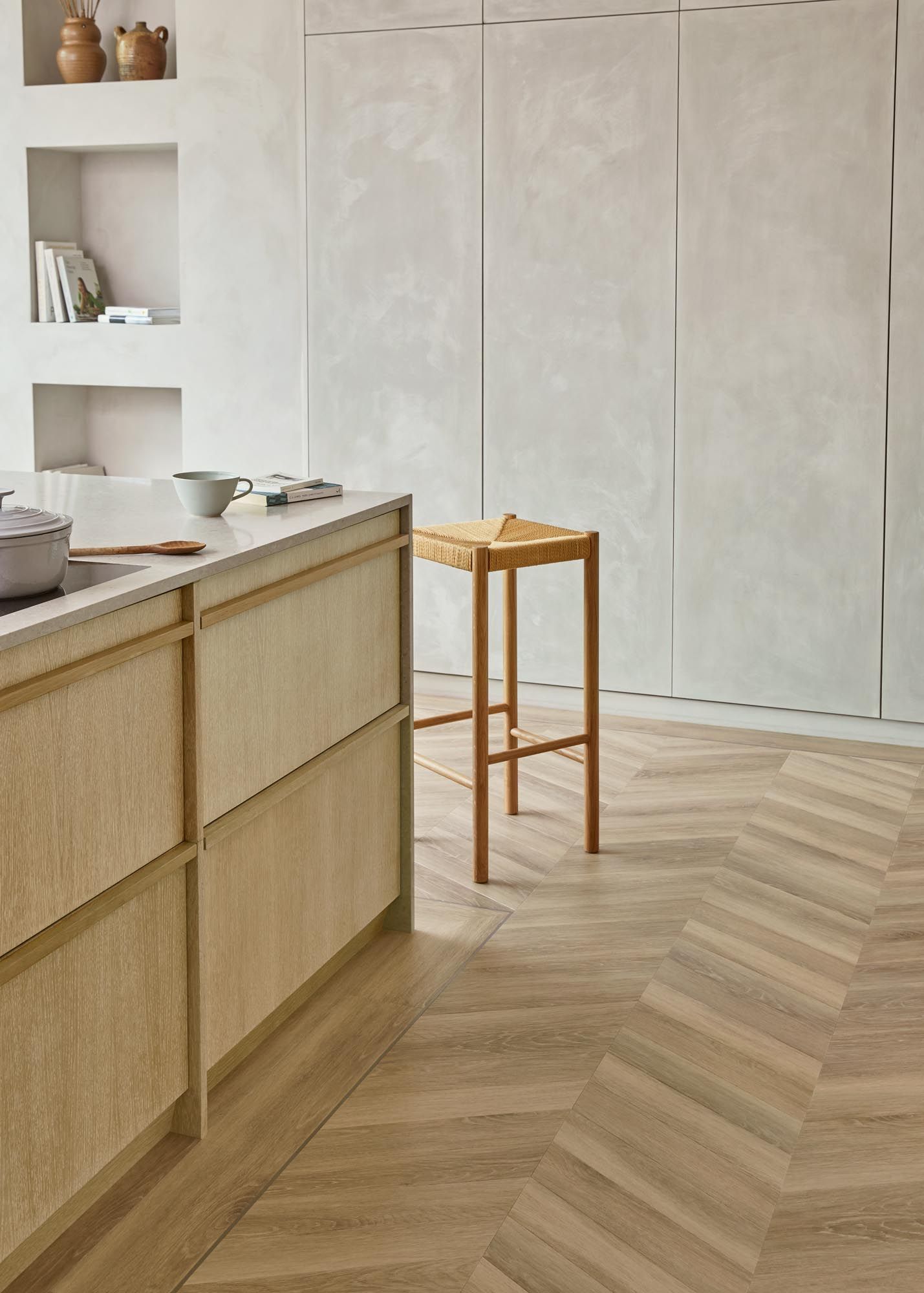 Küche mit hellen Holzschränken, weißen Wänden und Fischgrätparkett. Ein Hocker steht an der Kücheninsel.