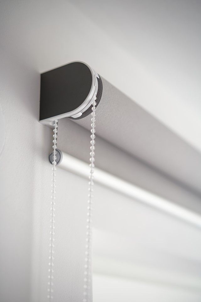Nahaufnahme eines grauen Rollos an einer weißen Wand. Die Zugschnur hängt herunter.