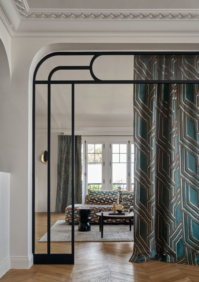 Ein schwarz umrahmter Türrahmen mit türkis-gold gemusterten Vorhängen führt in ein Wohnzimmer mit einem gemusterten Sofa.