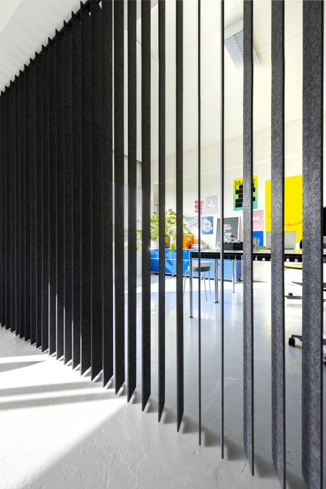 Dunkelgraue, vertikale Lamellen verdunkeln teilweise einen hell erleuchteten Büroraum.