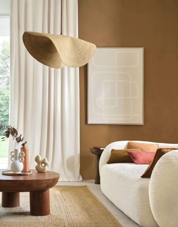 Gemütliches Wohnzimmer mit beigem Sofa, braunen Wänden und moderner Kunst. Darüber hängt eine geflochtene Lampe.