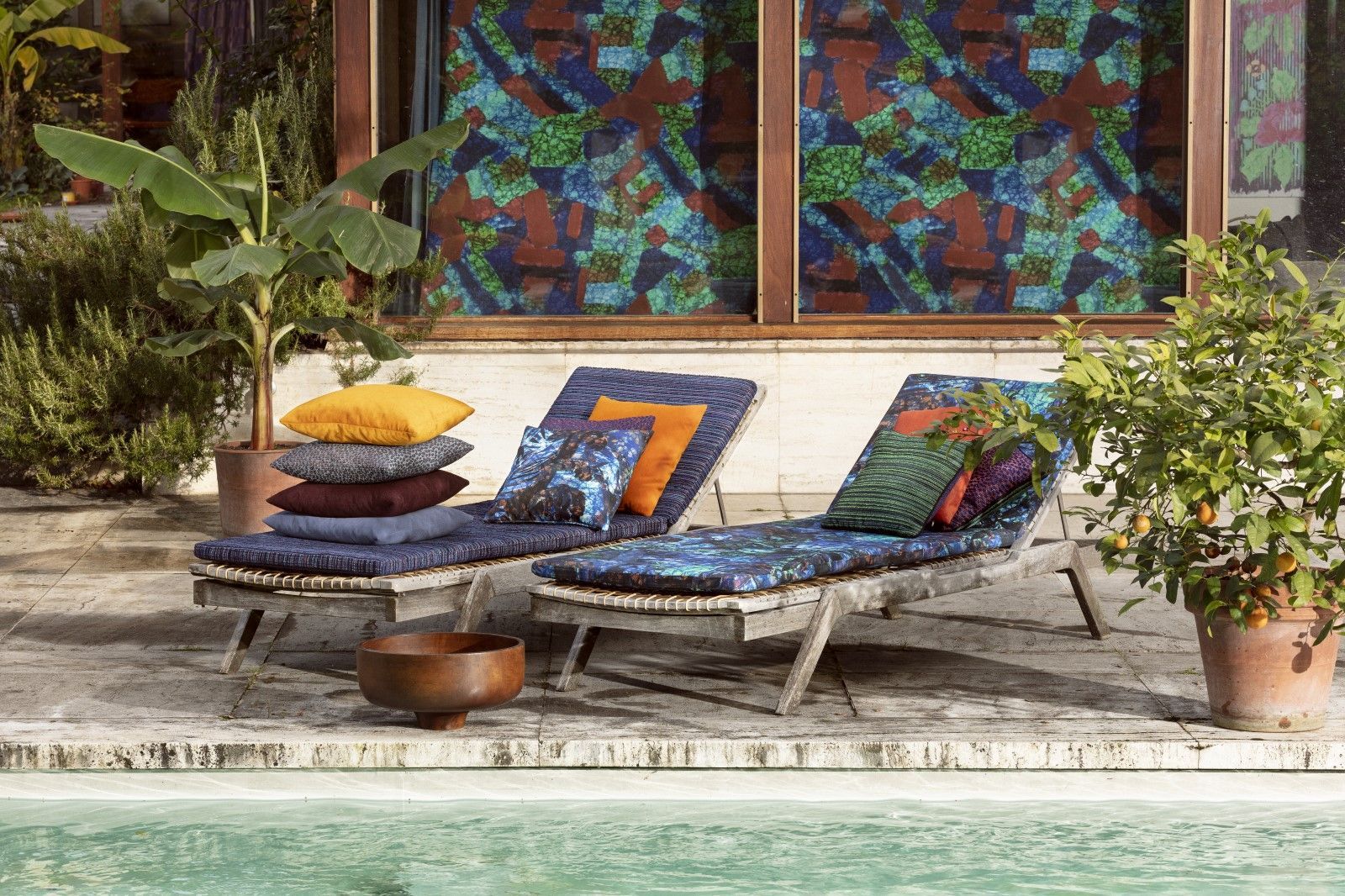 Zwei Liegestühle am Pool, gemusterte Kissen, Pflanzen und ein gemustertes Fenster.