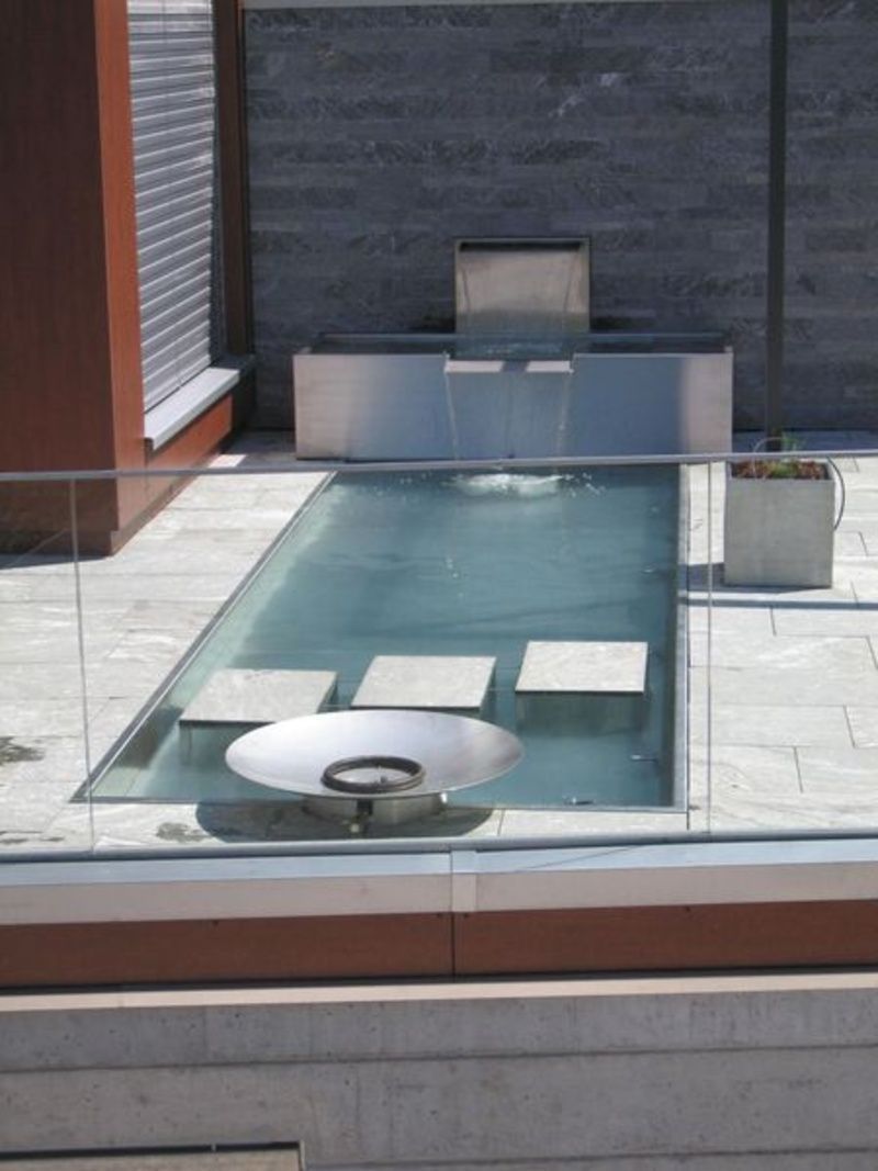 Ein Glaszaun umgibt einen Swimmingpool mit einem Springbrunnen im Hintergrund. Foto von der Artmetal GmbH