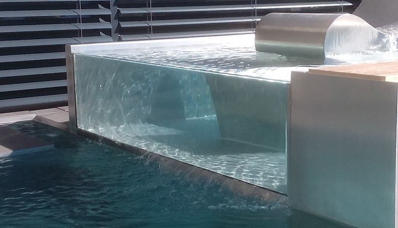 Ein Swimmingpool aus Edelstahl mit einer klaren Glaswand und einem Wasserfall. Foto von der Artmetal GmbH
