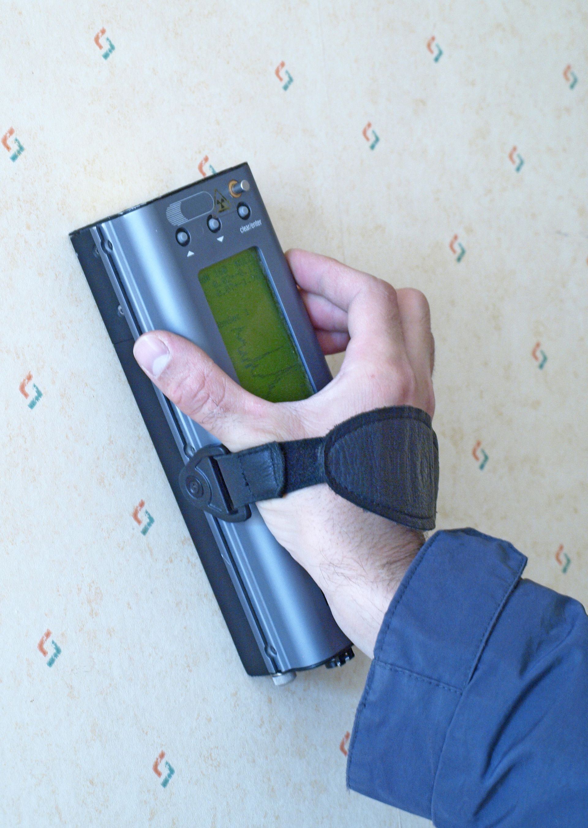 Main tenant un détecteur de montants muraux, attaché au poignet, contre un mur à motifs.