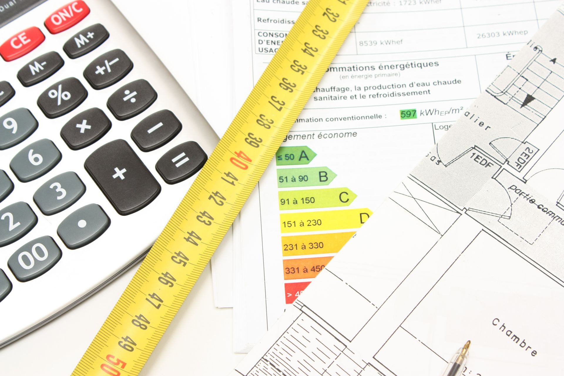 Calculatrice, ruban à mesurer et documents avec tableau d'évaluation énergétique et plan architectural sur une surface blanche.