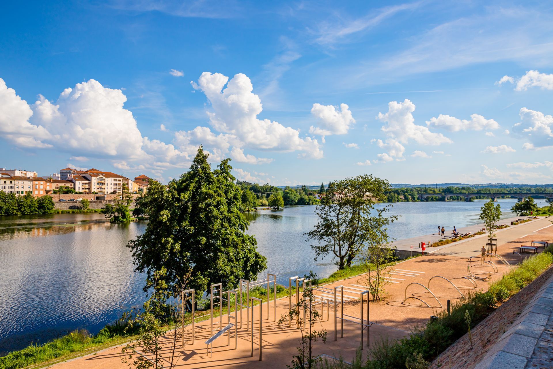 Vue panoramique d'une rivière avec des arbres, un sentier au bord de l'eau et des bâtiments sous un ciel bleu avec des nuages ​​​​duveteux.