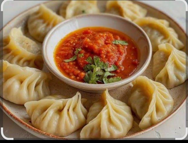 Momos al vapor dispuestos en un plato alrededor de un tazón de salsa roja, adornados con cilantro.